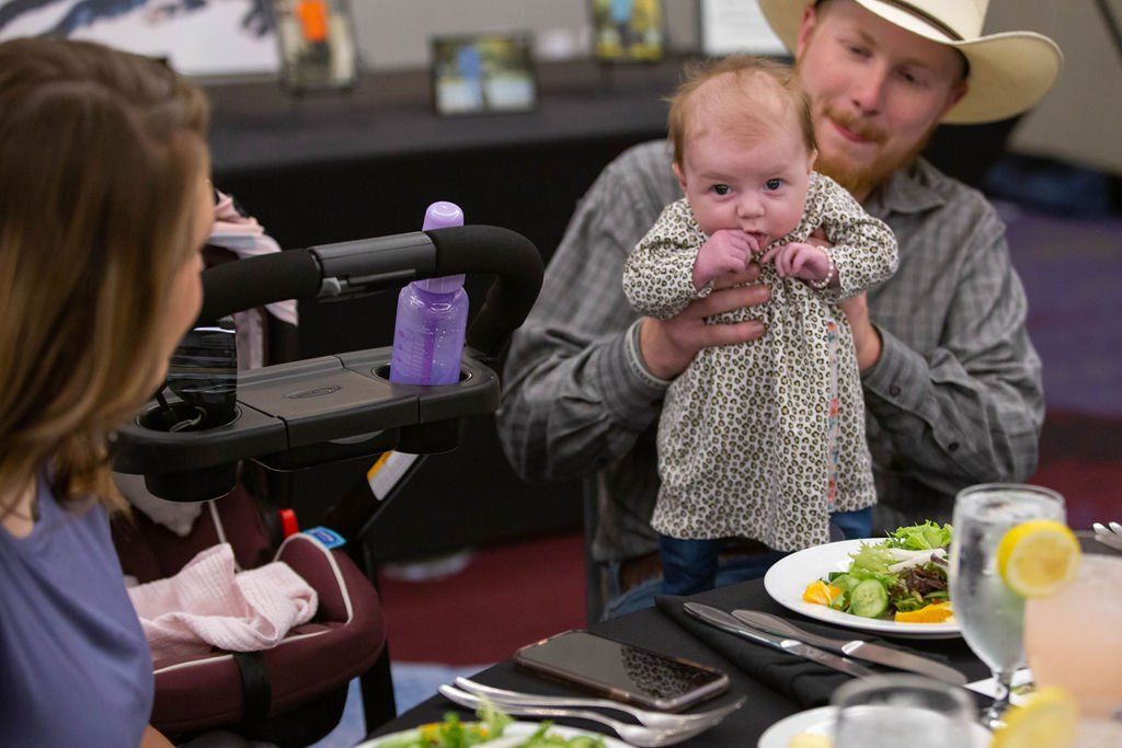 A person wearing a cowboy hat holds an infant at a dinner table while sitting across from another person.