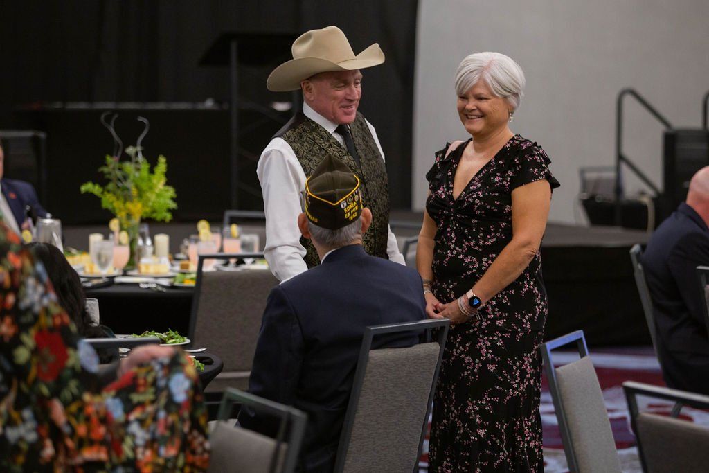 Three people converse in a conference room; one man wears a cowboy hat and patterned vest, another wears a crown cap.