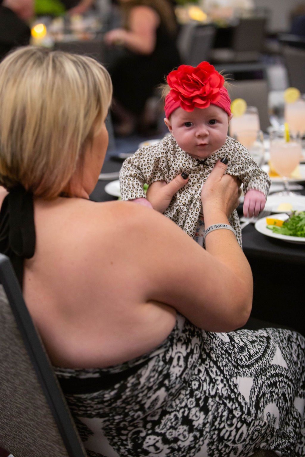 A person in a black and white patterned dress holds a baby wearing a red rose headband at a dining table.