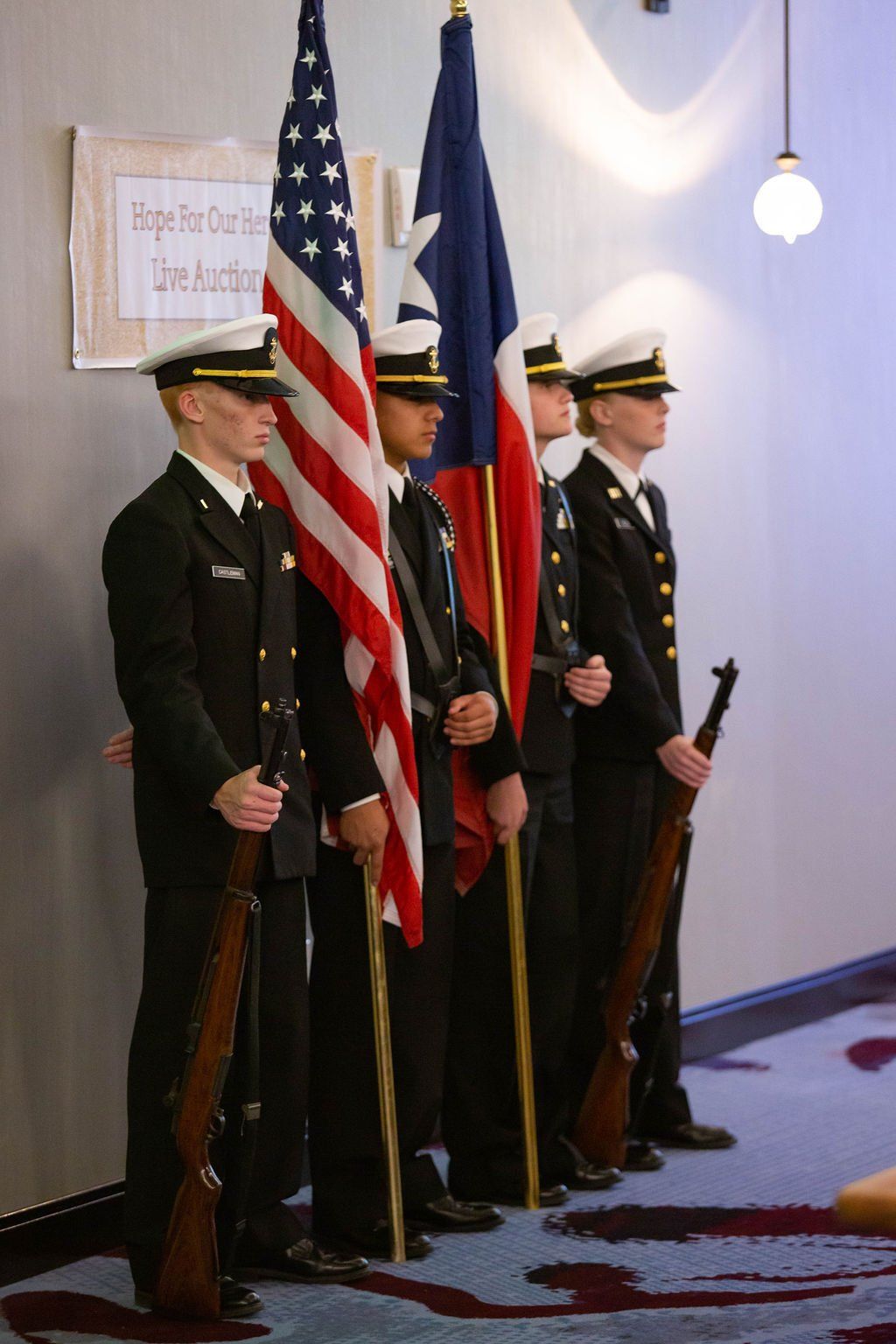 Four honor guard members in dark formal uniforms and hats hold flags and rifles while standing in a line indoors.