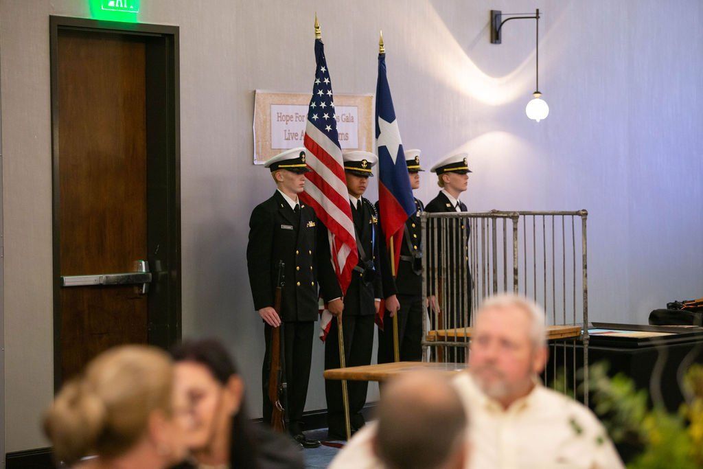 A color guard in dress uniforms stands at attention holding the US and Texas flags in an indoor room with event guests.