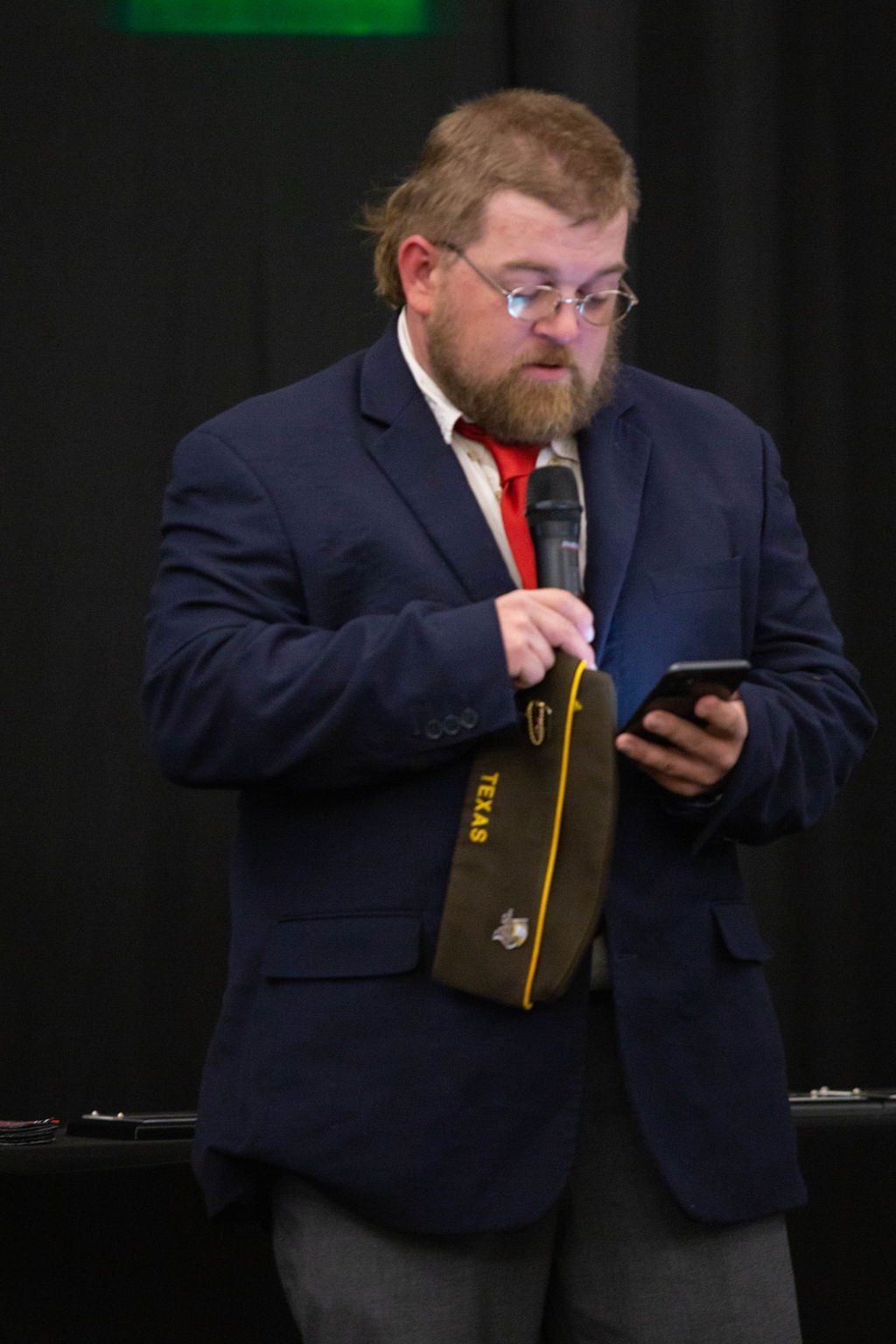A man in a navy blazer and red tie holds a microphone and a smartphone while wearing a tan Texas cap over his arm.