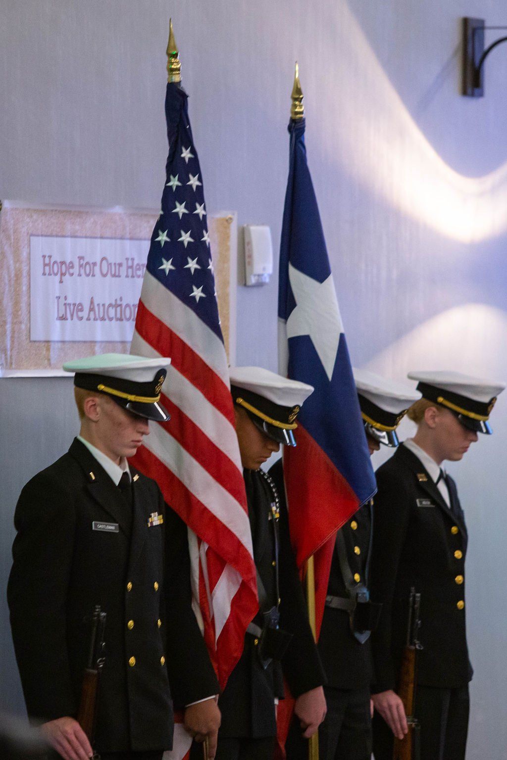 Four members of a color guard in dark dress uniforms holding American and Texas flags during an indoor ceremony.