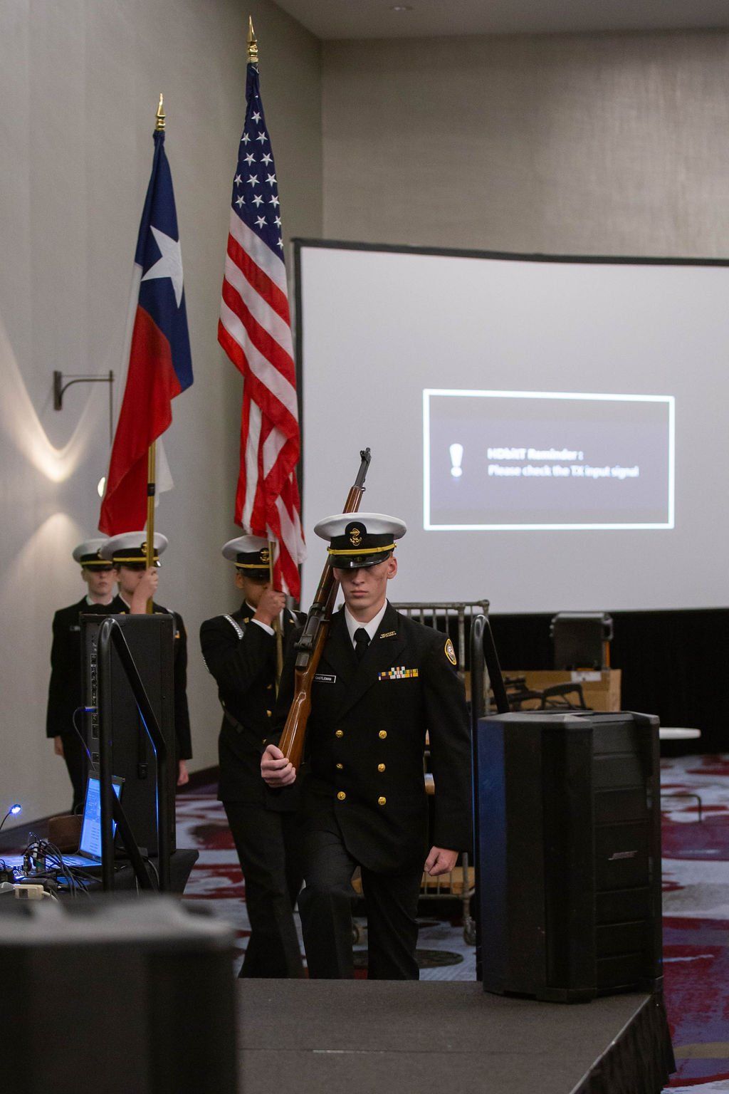 A color guard in navy uniforms marches with rifles past the Texas and U.S. flags in a room with a projector screen.