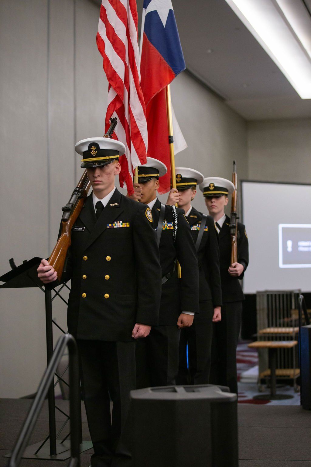 A color guard in Navy uniforms carries the U.S. and Texas flags and rifles in a formal indoor ceremony.