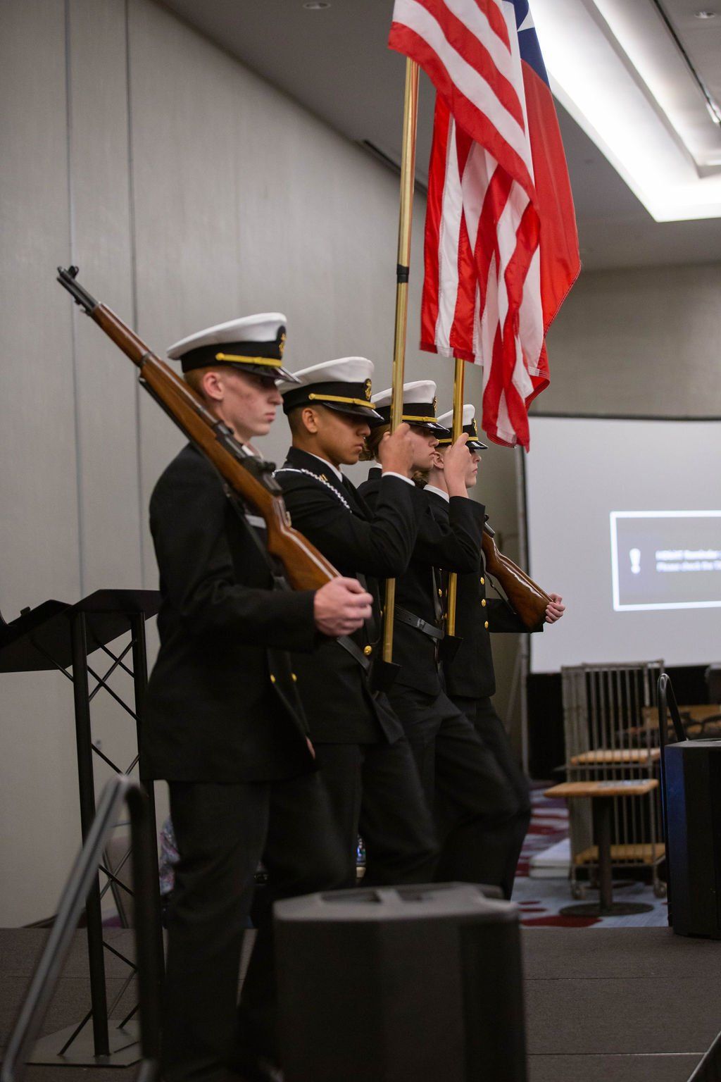 Four uniformed color guard members in formal military attire stand in a row holding rifles and an American flag.