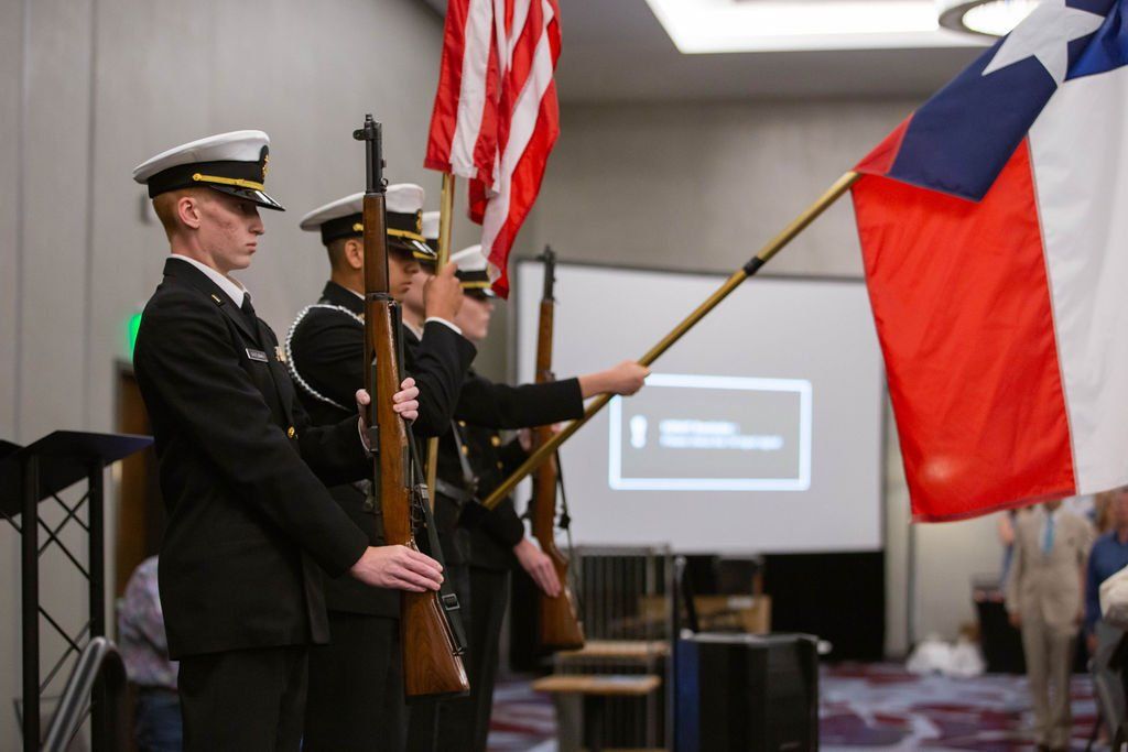 Color guard in formal uniforms standing in a room, holding the U.S. and Texas flags and rifles in a presentation formation.