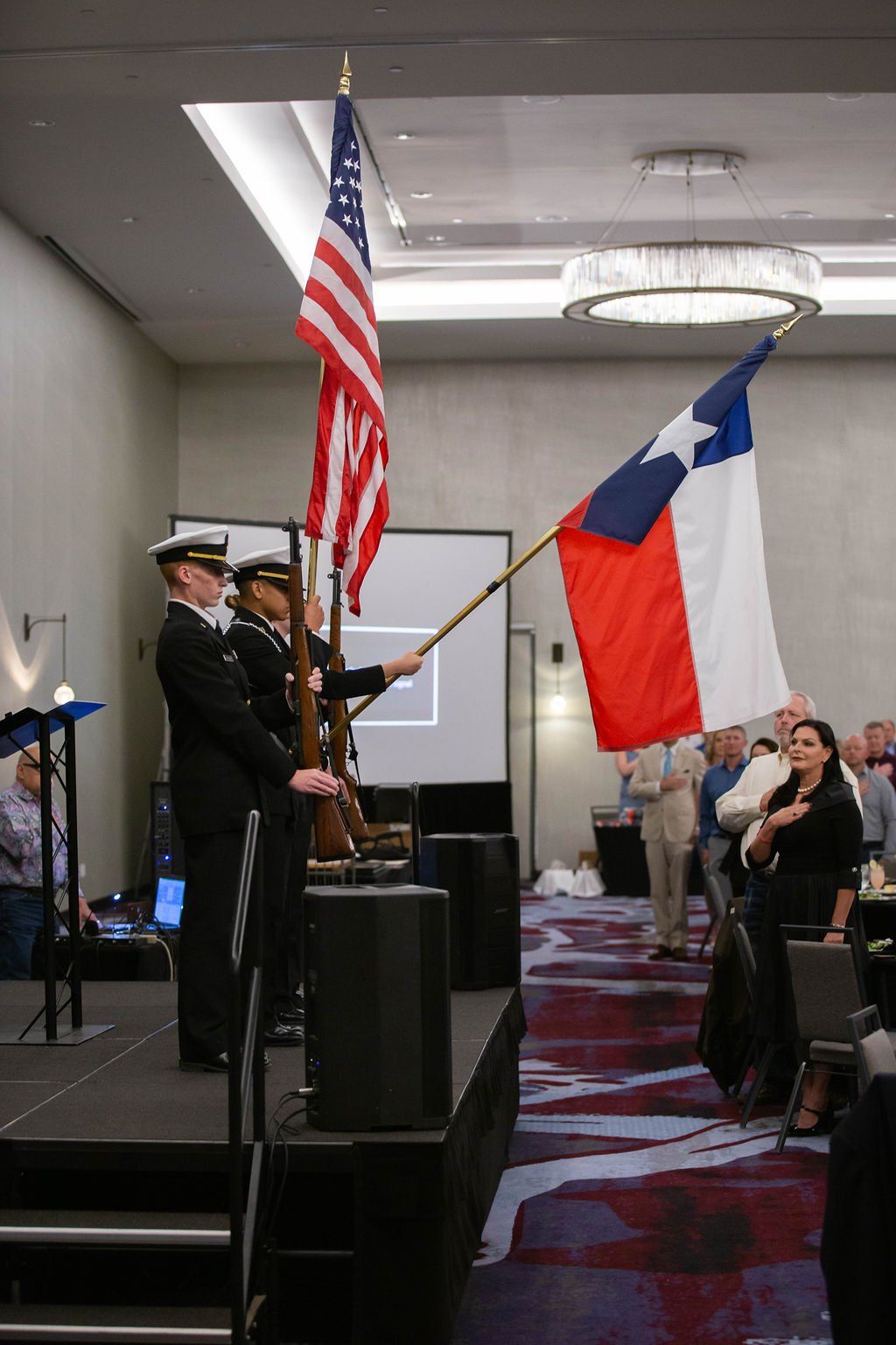 Two service members in uniform stand on a stage holding the US and Texas flags during an indoor event.
