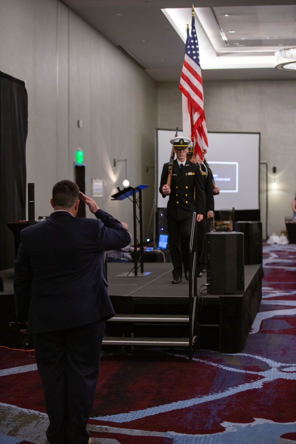 A person in a suit salutes as a color guard in dress blues carries the American flag across an indoor stage.