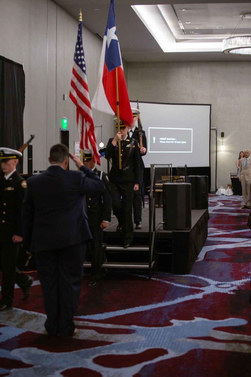 A color guard in military dress uniforms marches across a conference room stage, carrying the US and Texas flags.
