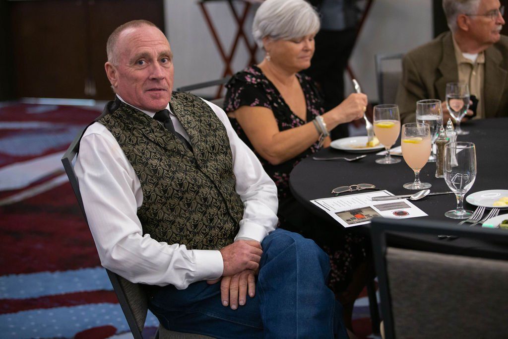 A man in a patterned vest sits at a table during a dinner event, while others eat in the background.
