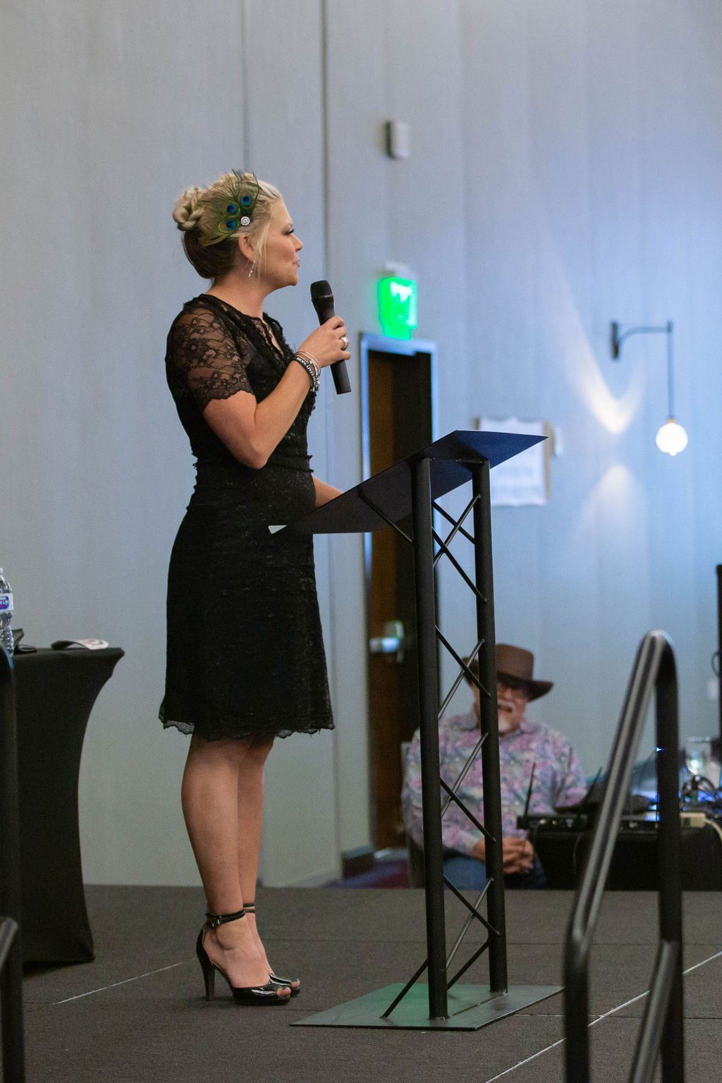 A person in a black dress stands at a podium on a stage, speaking into a microphone with someone seated in the background.