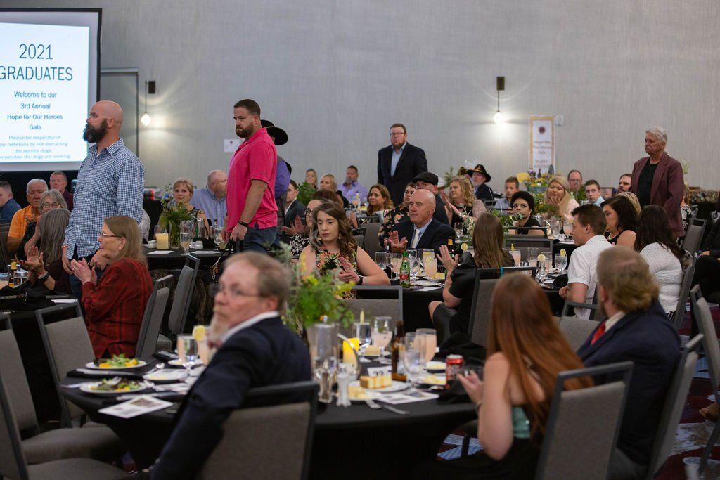 A group of people seated at tables in a large hall during an event, with a screen in the background displaying 2021 text.