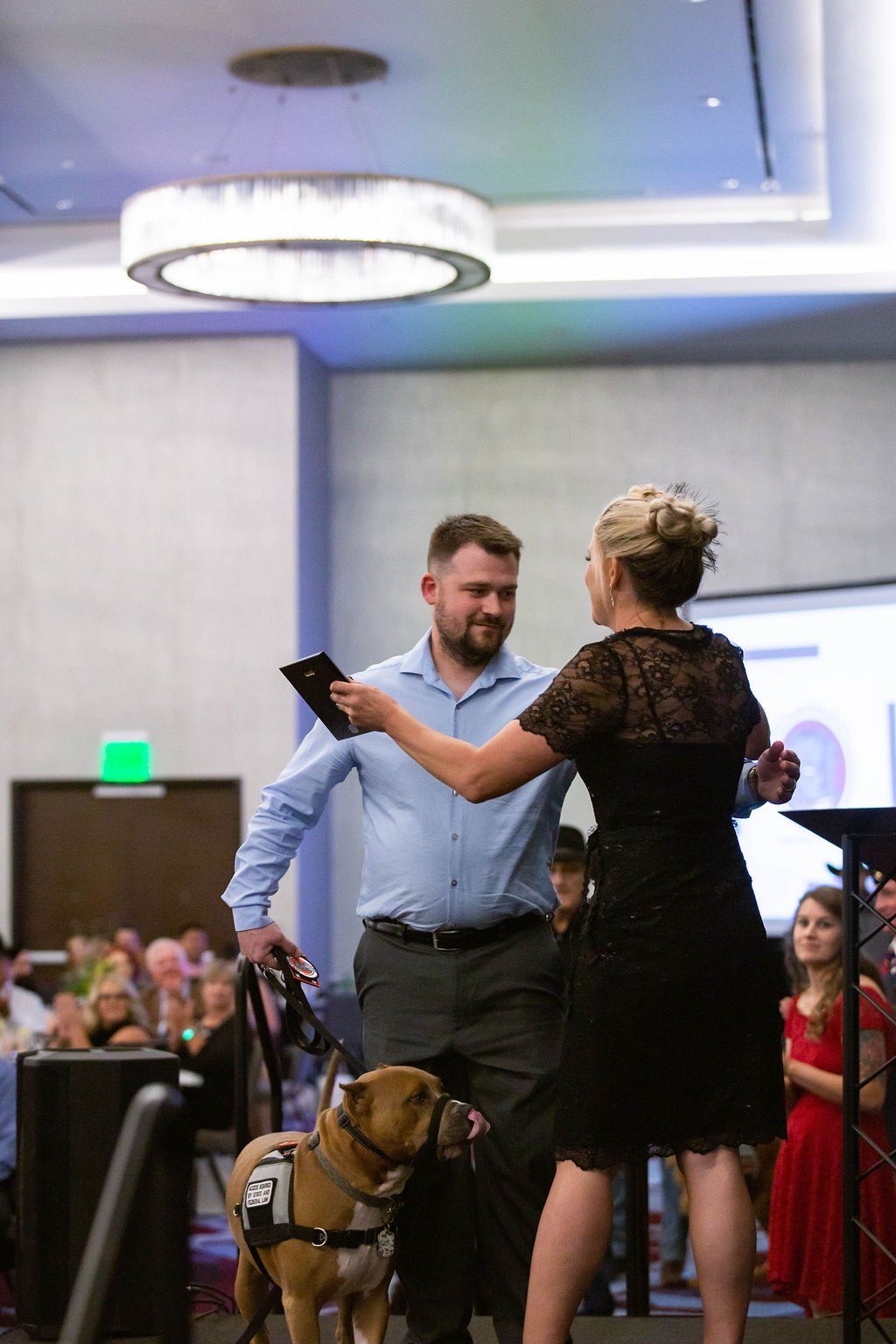 A man with a service dog and a woman in a black dress stand together on a stage at an indoor event.