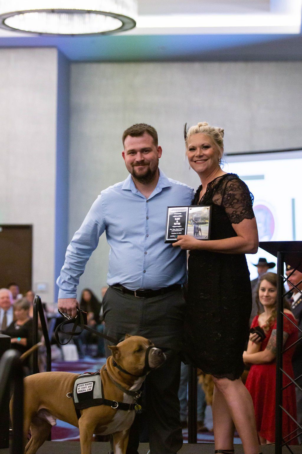 A man and woman holding an award stand indoors with a tan service dog wearing a harness.