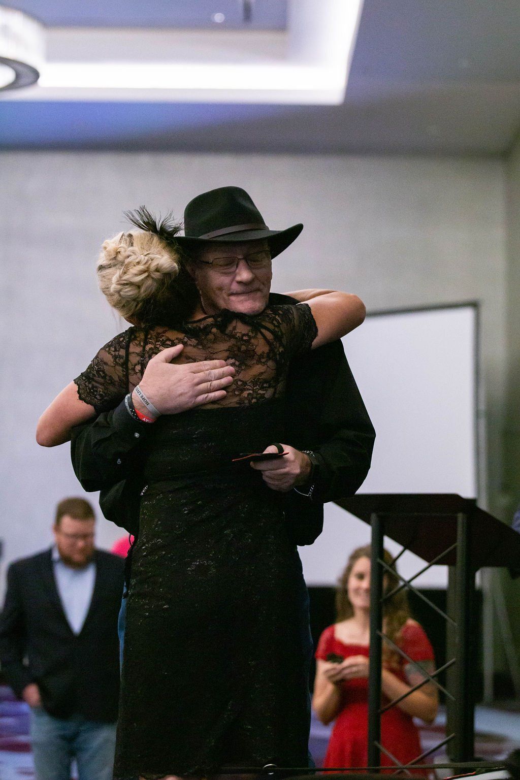 A person in a black hat and black clothes embraces another person in a black lace dress at an indoor event.