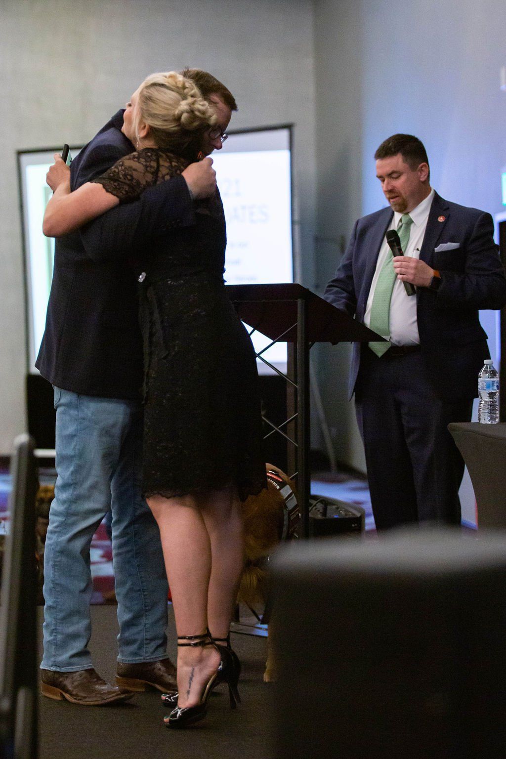 A woman and man in formal attire embrace on a stage, while a man in a suit speaks into a microphone at a podium nearby.