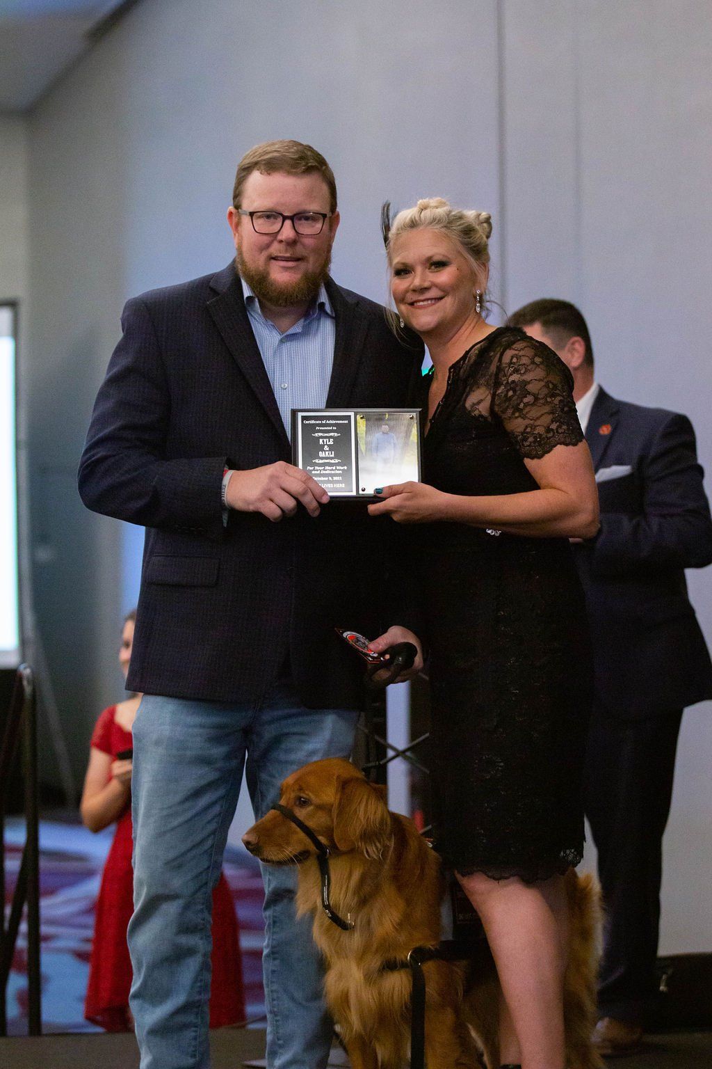 A man and woman standing together holding a plaque with a golden retriever at their feet in an indoor event setting.