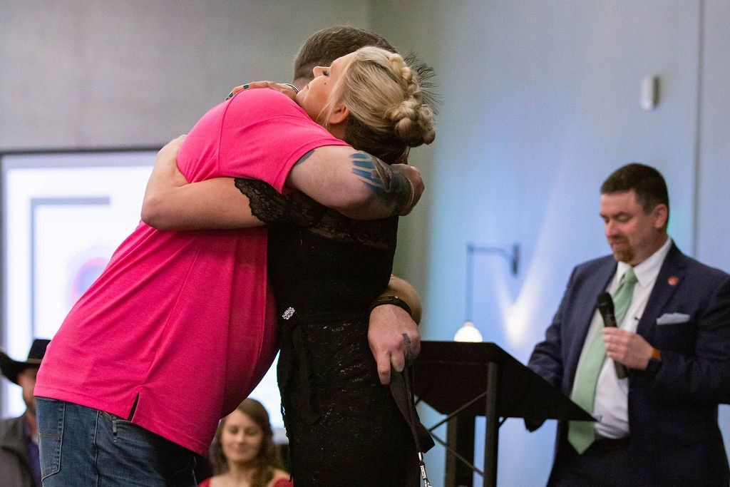 Two people embrace in an indoor setting, while a person in a suit stands nearby holding a microphone at a podium.