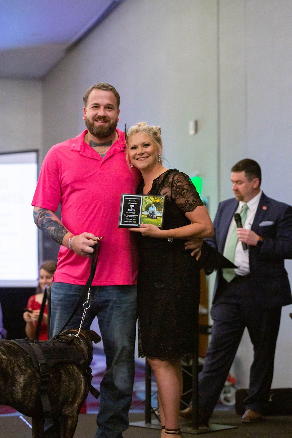 A man in a pink shirt and a woman in a black dress hold an award together, standing next to a dog on a leash on stage.