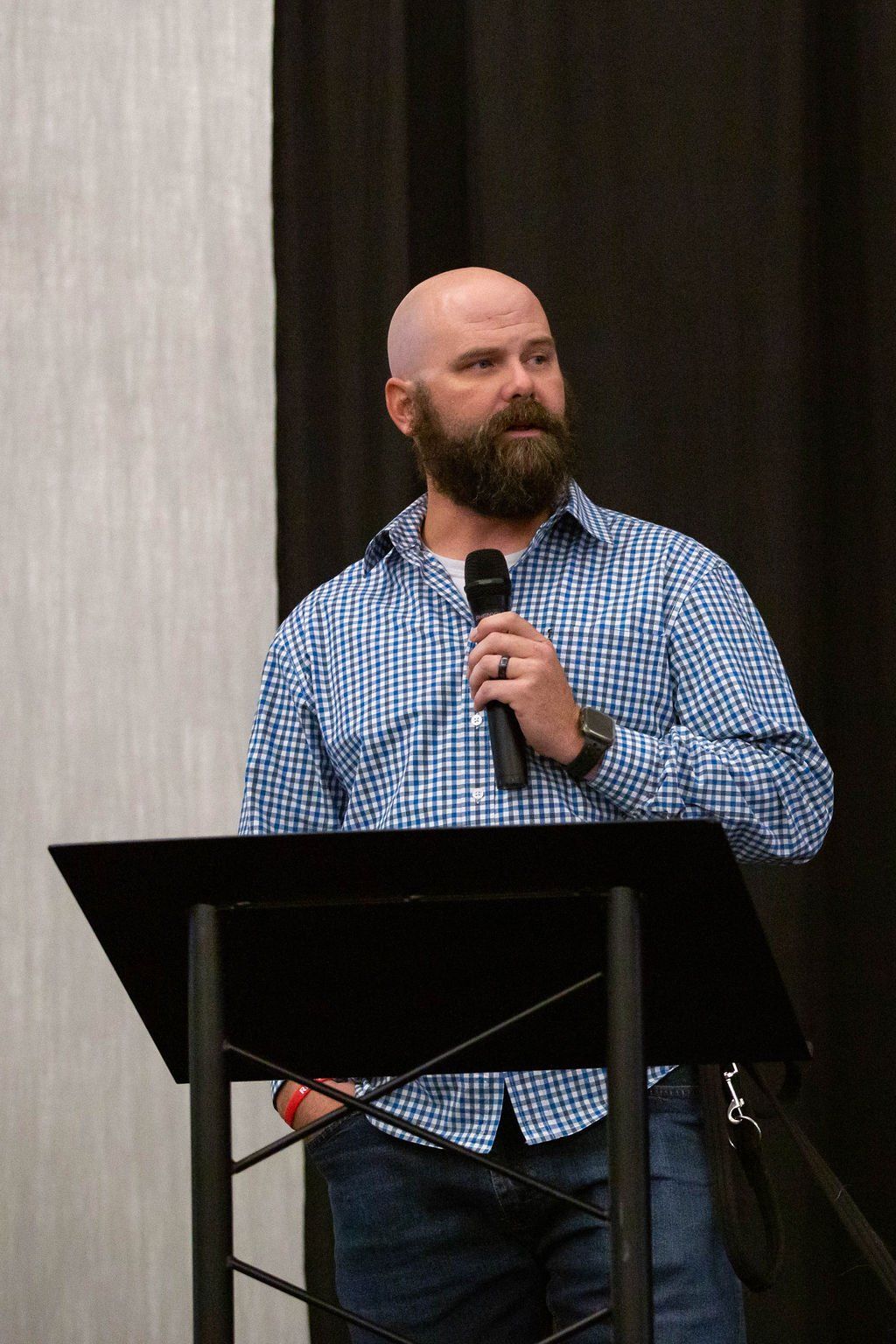 A bald person with a beard wearing a blue patterned button-down shirt speaks into a microphone at a black podium.