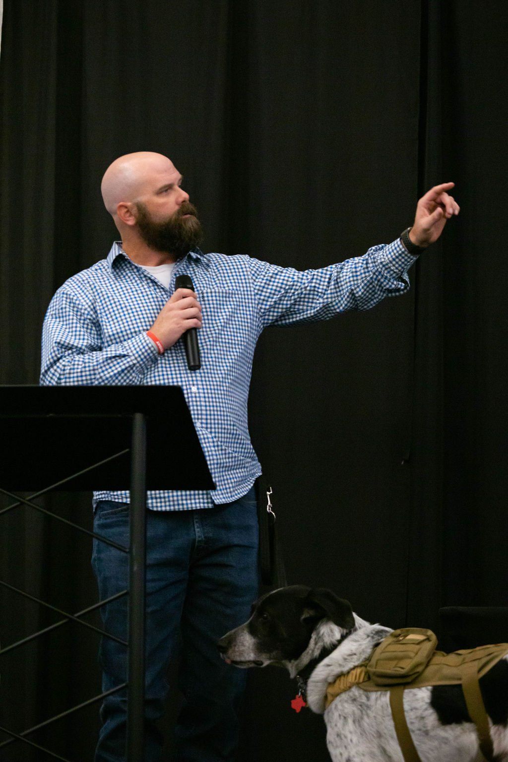 A man with a beard and patterned shirt stands at a podium, speaking into a microphone and pointing, with a dog nearby.