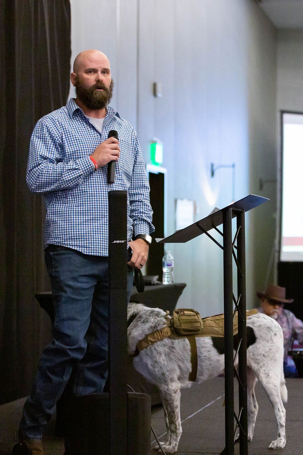 A bearded person stands at a podium speaking into a microphone, with a service dog wearing a tan harness nearby.