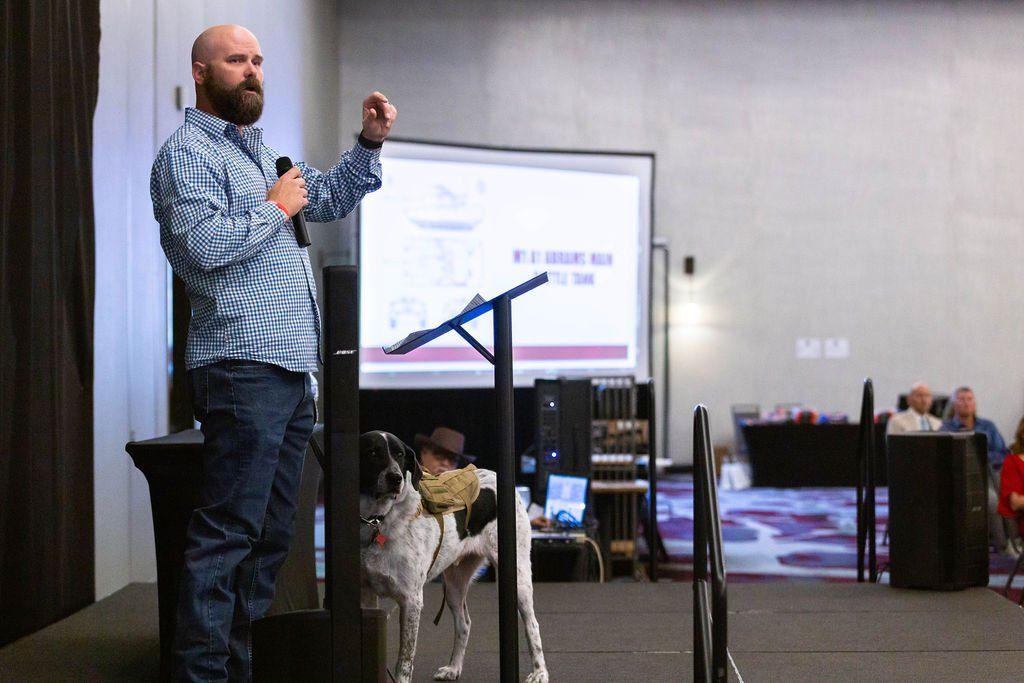 A speaker stands on a stage next to a spotted dog, gesturing while presenting to an audience in a conference room.