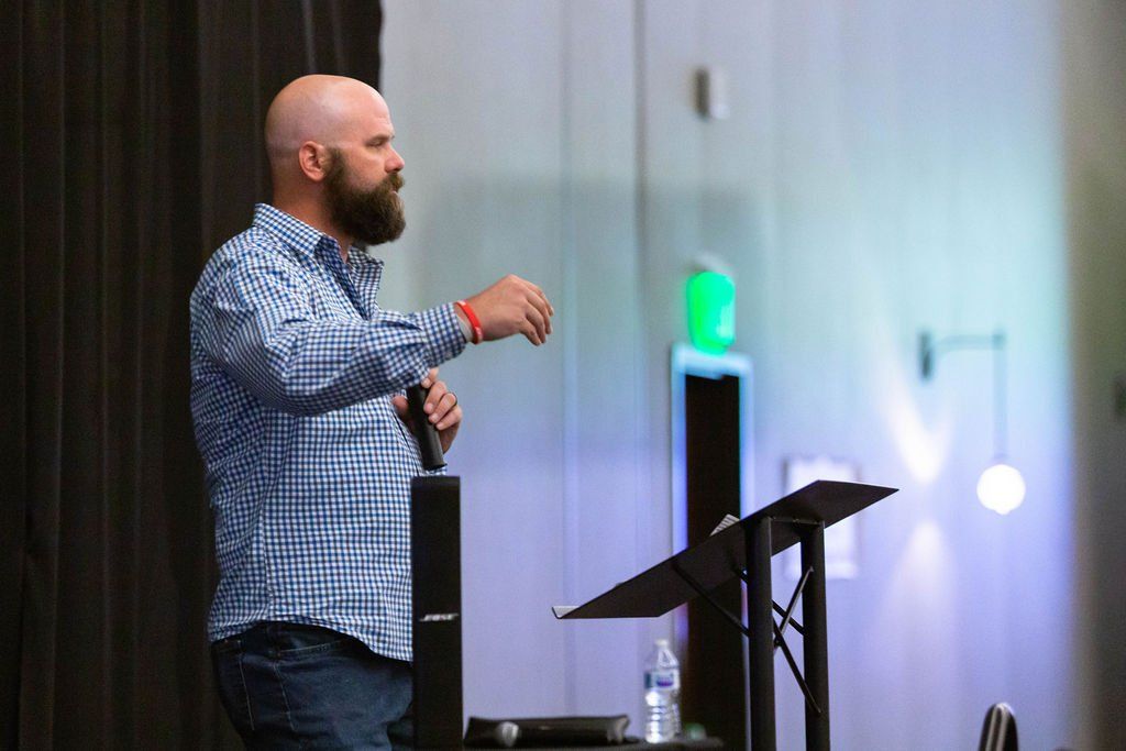 A speaker with a beard wearing a plaid shirt gestures while standing at a podium in a dimly lit room with blue lighting.