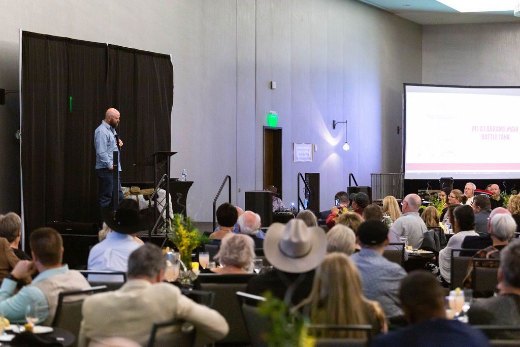 A presenter standing on a stage at a conference, speaking to a large audience seated at tables in a dimly lit ballroom.