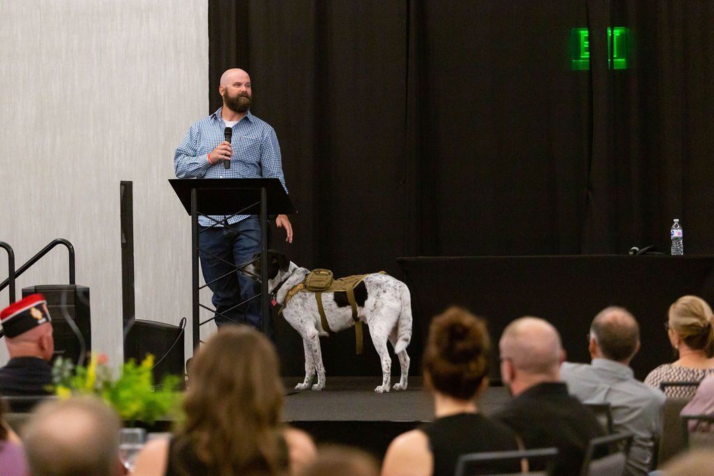 A speaker stands at a podium giving a presentation with a service dog wearing a tactical vest standing beside them.