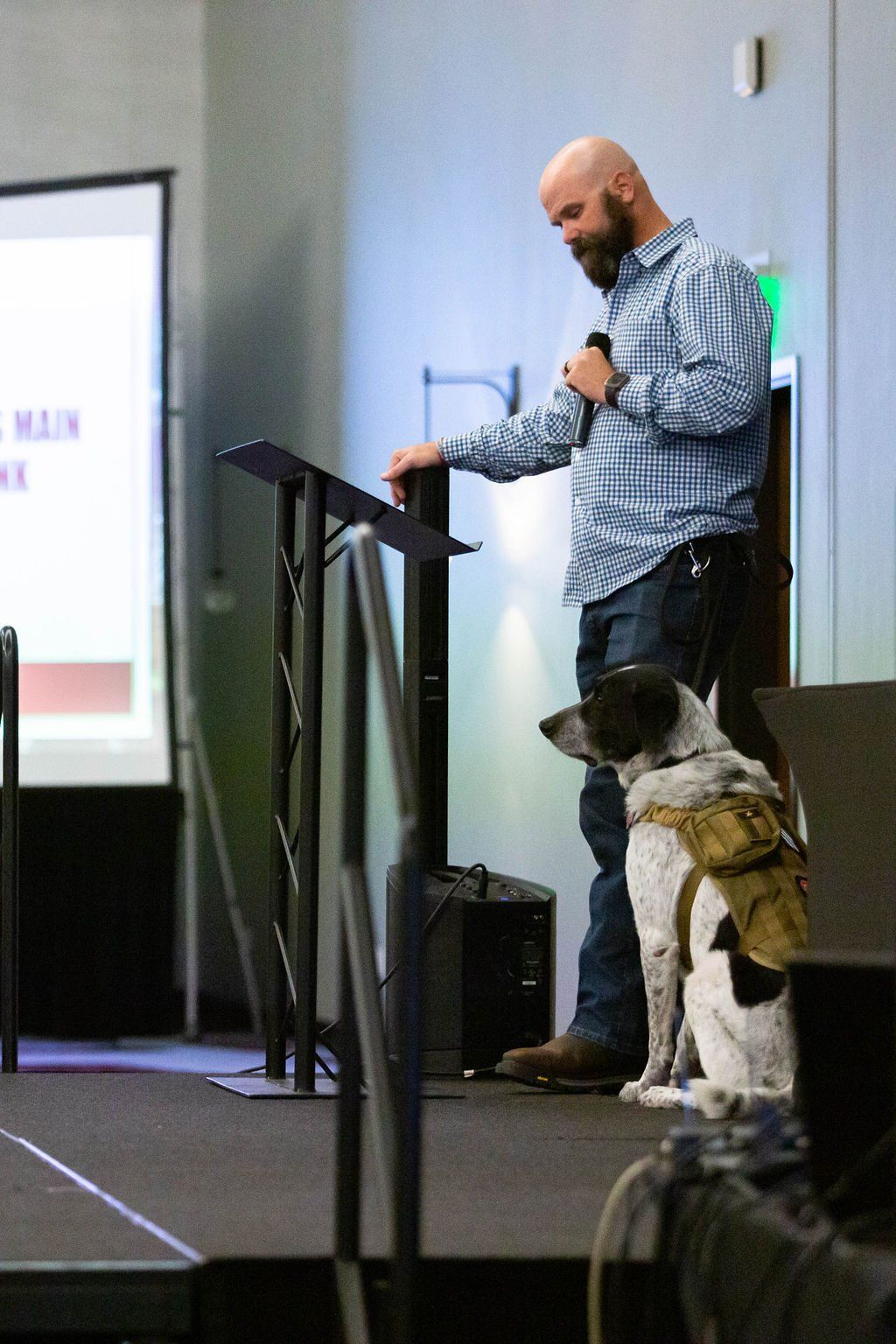 A bearded speaker stands at a podium next to a service dog wearing a tan tactical vest on a stage.