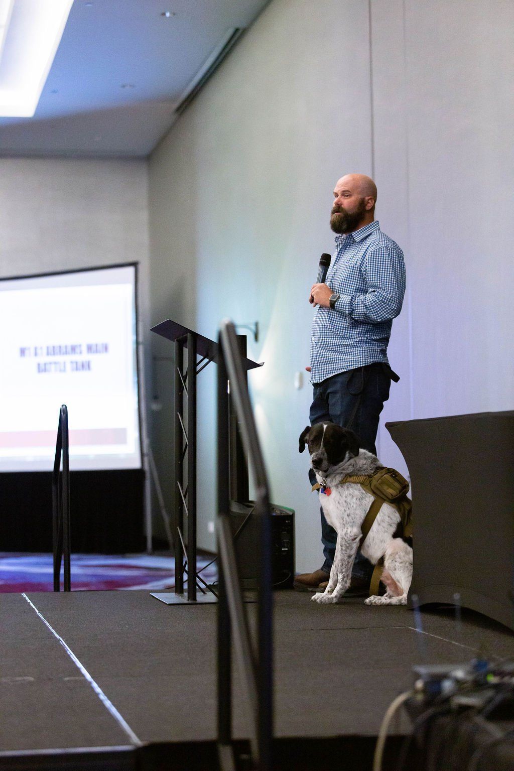 A man stands at a lectern on a stage holding a microphone, with a dog wearing a tactical vest sitting calmly beside him.