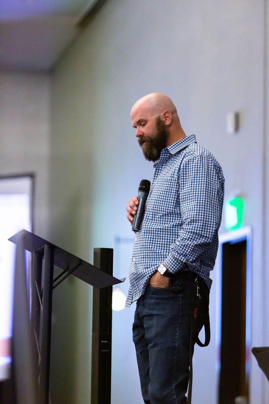 A bearded person in a blue checkered shirt stands at a podium holding a microphone, looking down at notes.
