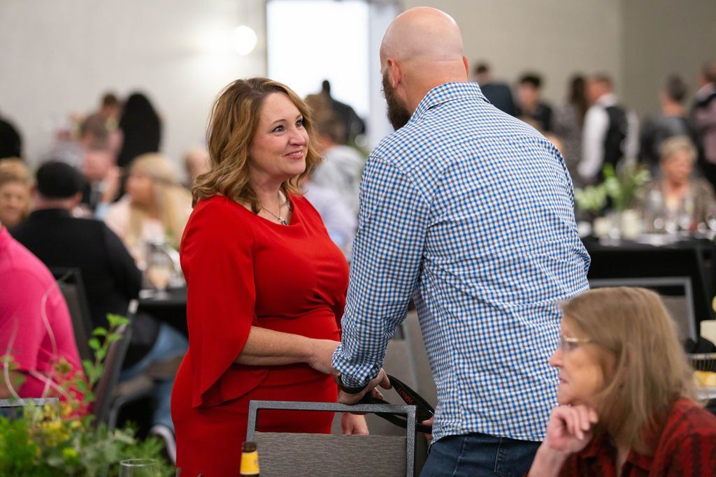 A woman in a red dress smiles while speaking to a man in a blue button-down shirt at a crowded event.