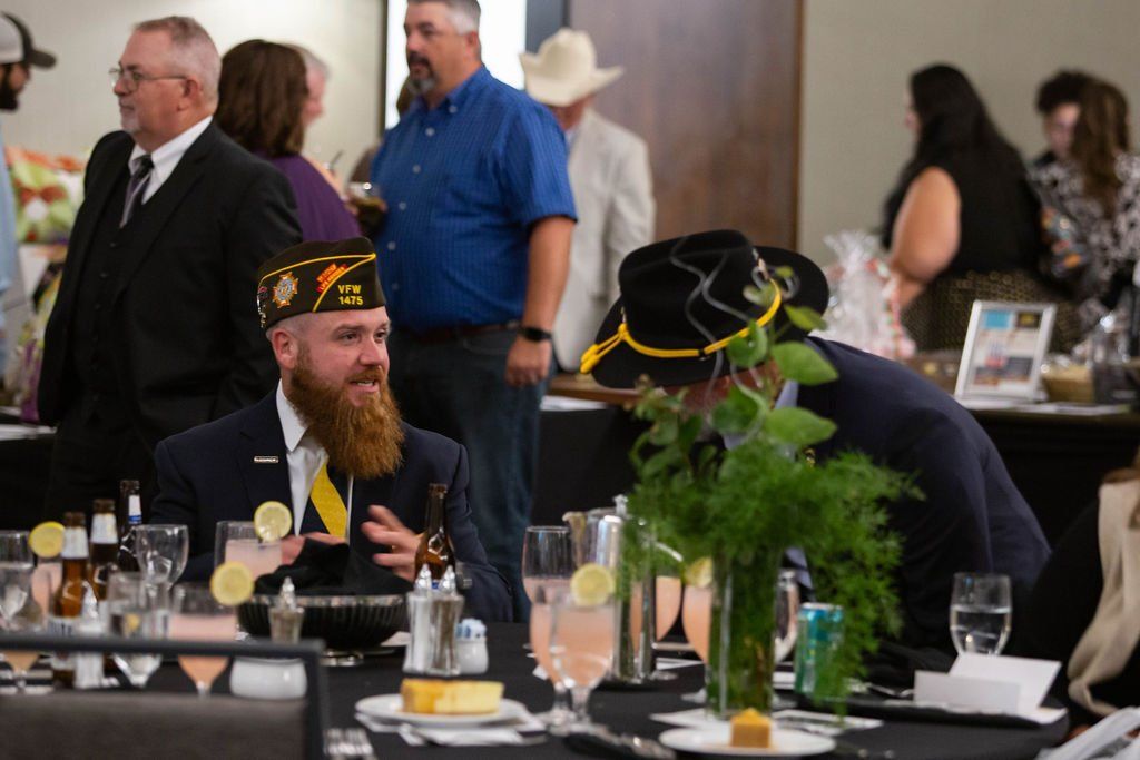 People in formal attire gather at tables during an indoor event; a man in a veteran’s cap sits in the foreground.