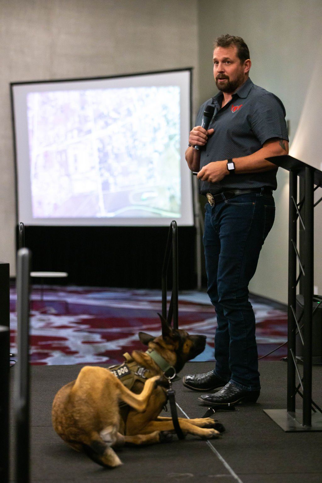 A person speaks into a microphone on stage in front of a projection screen as a service dog lies on the floor nearby.