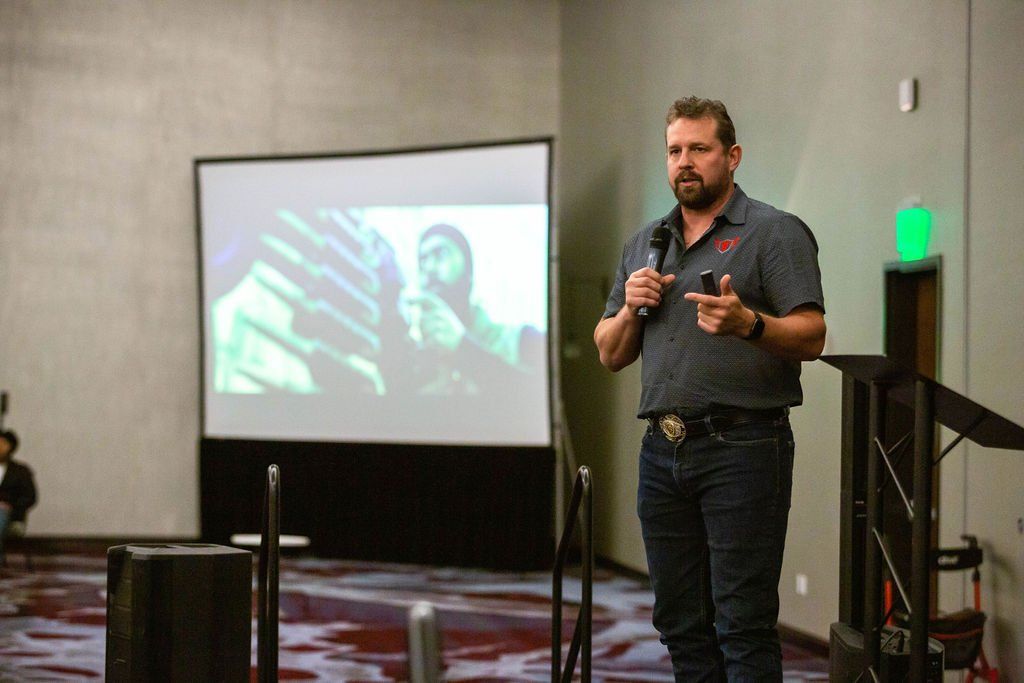 A speaker in a dark polo and jeans gestures while presenting in front of a projection screen in a modern conference room.