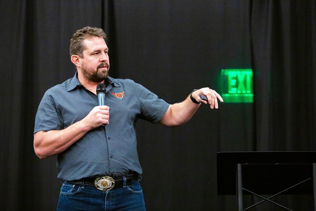 A speaker in a patterned shirt and jeans holds a microphone, gesturing toward an illuminated green exit sign.