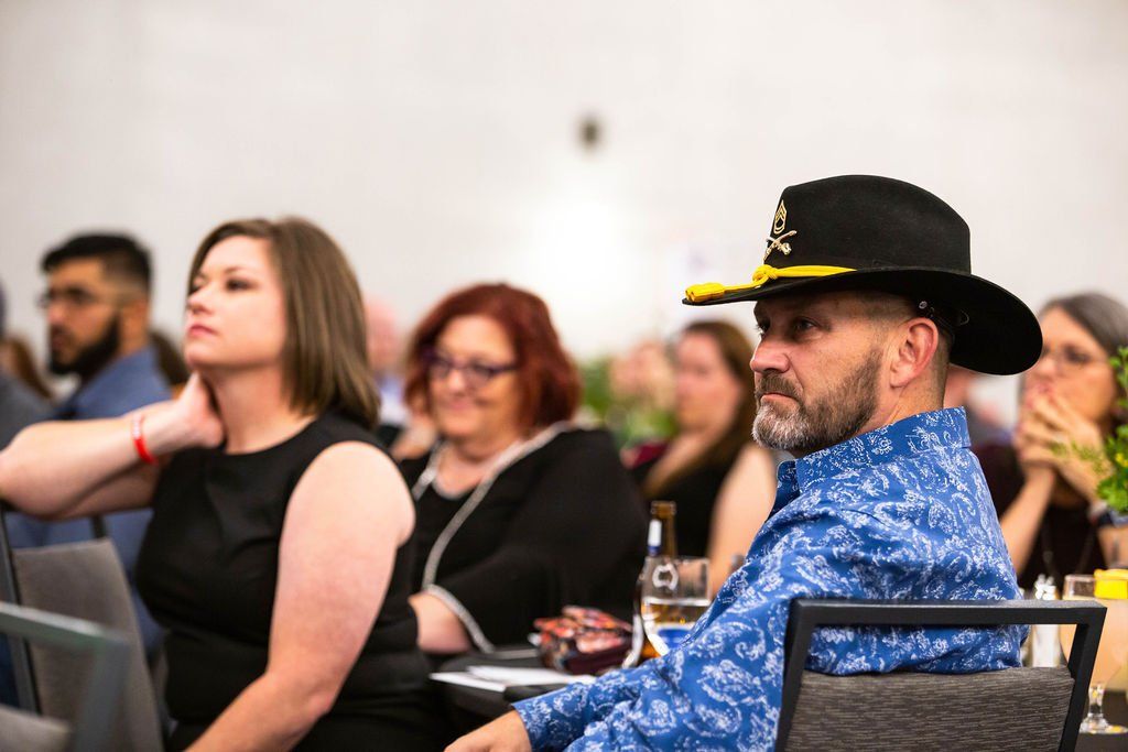 People seated at tables in a room, with a man in a black cowboy hat and a blue patterned shirt in the foreground.