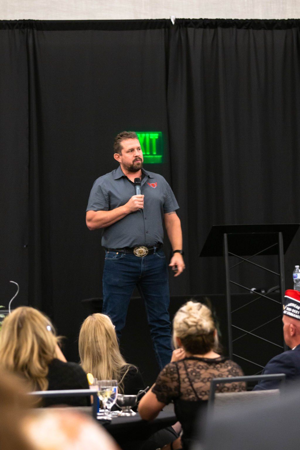 A man in a grey polo and jeans holds a microphone while speaking to an audience in front of a dark curtain.