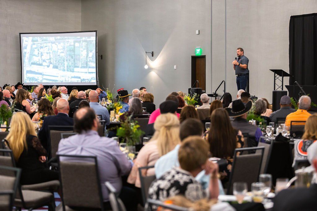 A speaker addresses a large audience gathered in a conference room with a large projection screen displaying an image.