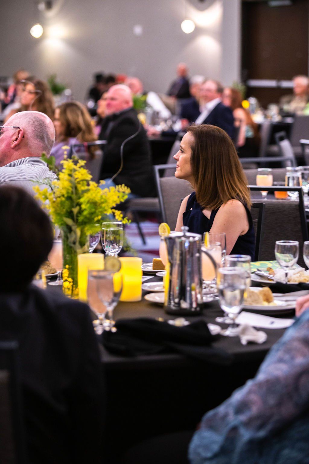 A banquet room with guests at round tables, focusing on a person seated at a table with yellow flowers and candles.