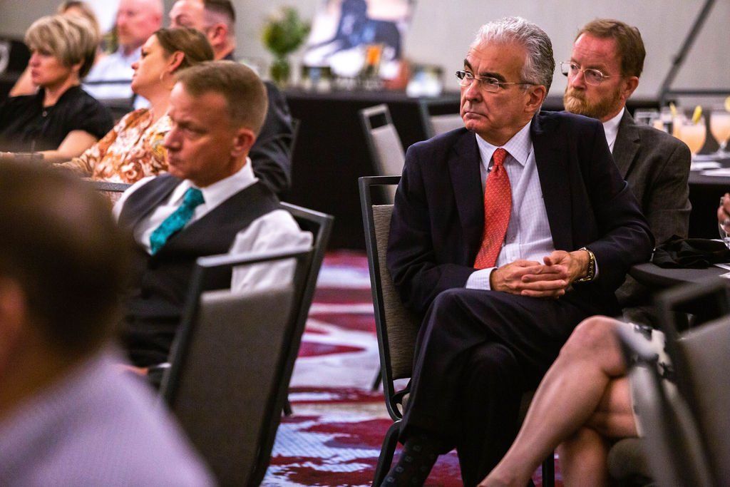 Audience members sit attentively at tables in a conference room, with a man in a suit and red tie in the foreground.