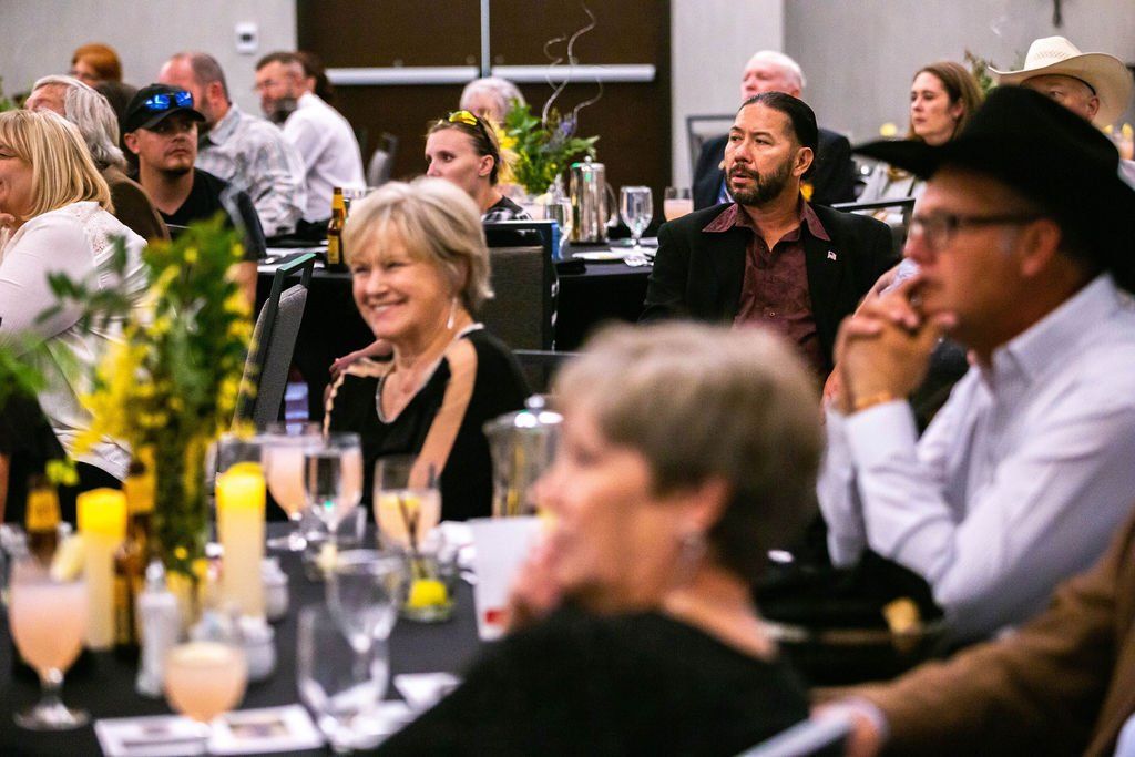 People sit at round tables in a conference room, listening to a presentation during an event.