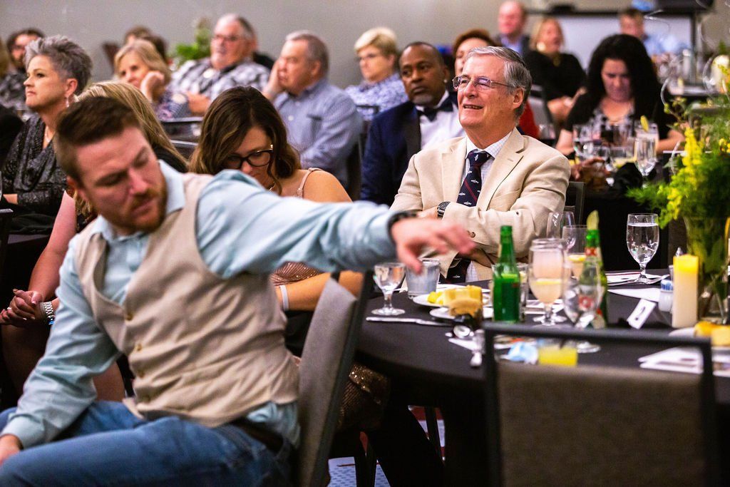 Attendees at a banquet hall, with a man in the foreground gesturing while sitting at a table with others.