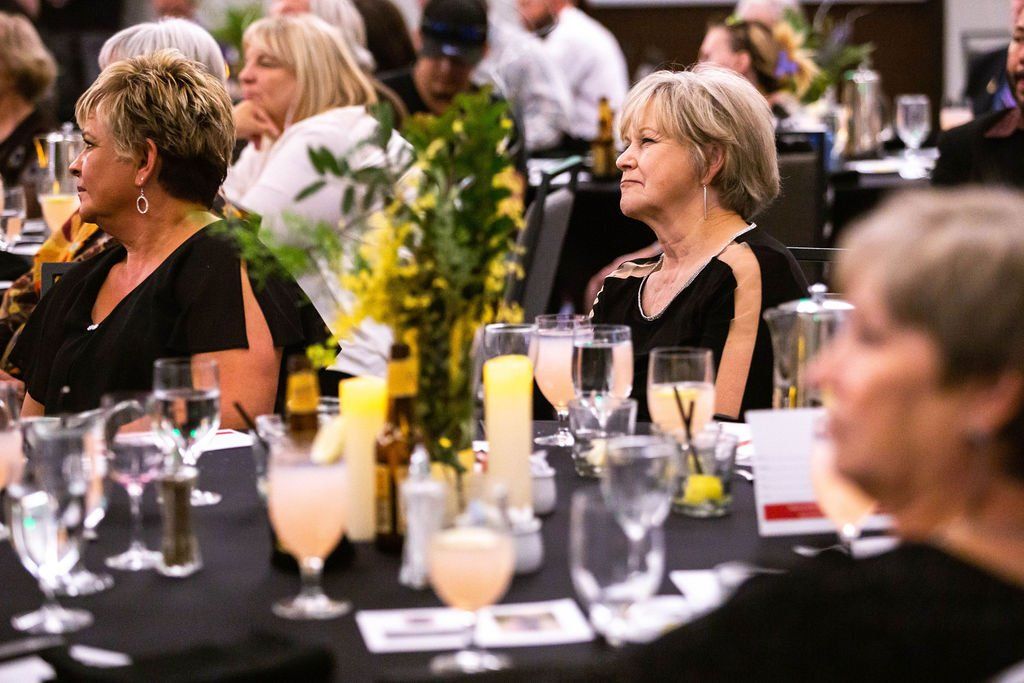 People sit at tables with black tablecloths and centerpieces at an indoor event, appearing to watch a presentation.