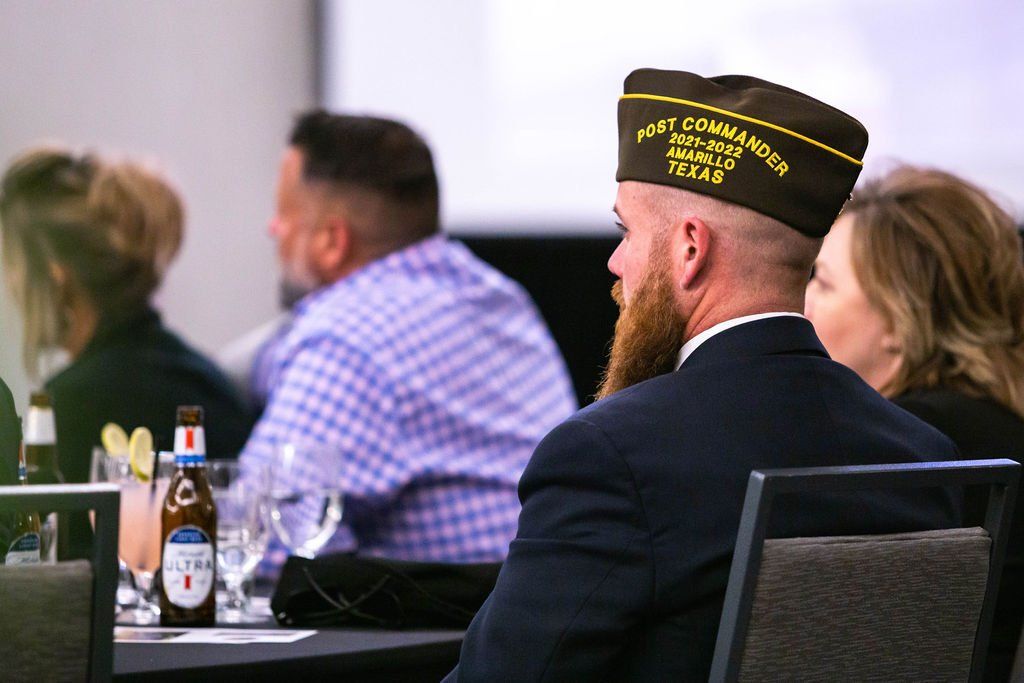 A person wearing a military-style cap sits at a table with drinks among others in a conference or meeting setting.