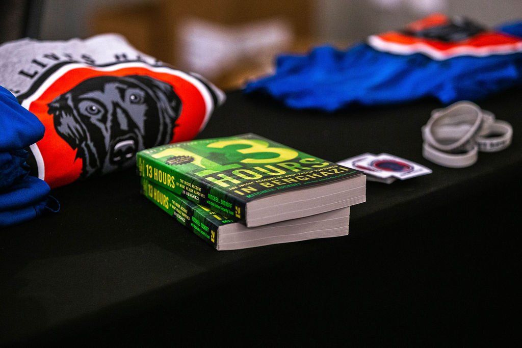 Two books with green covers stacked on a black table with shirts, wristbands, and a small decal nearby.
