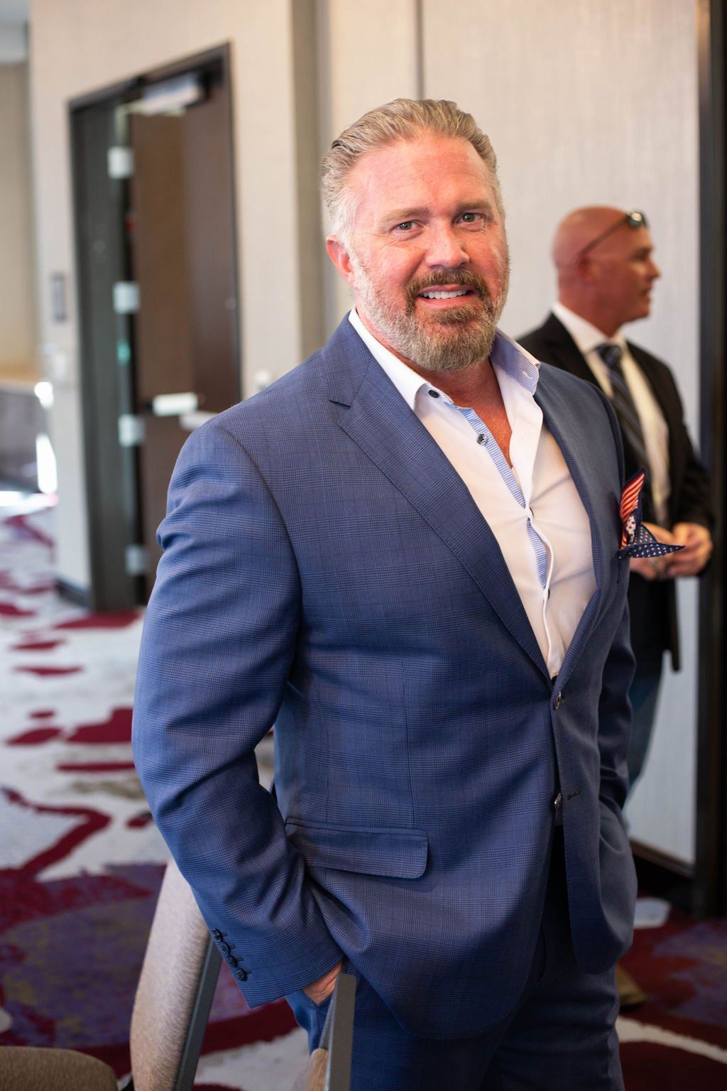 A person in a blue suit with a white shirt and pocket square smiles while standing in a room with patterned carpeting.